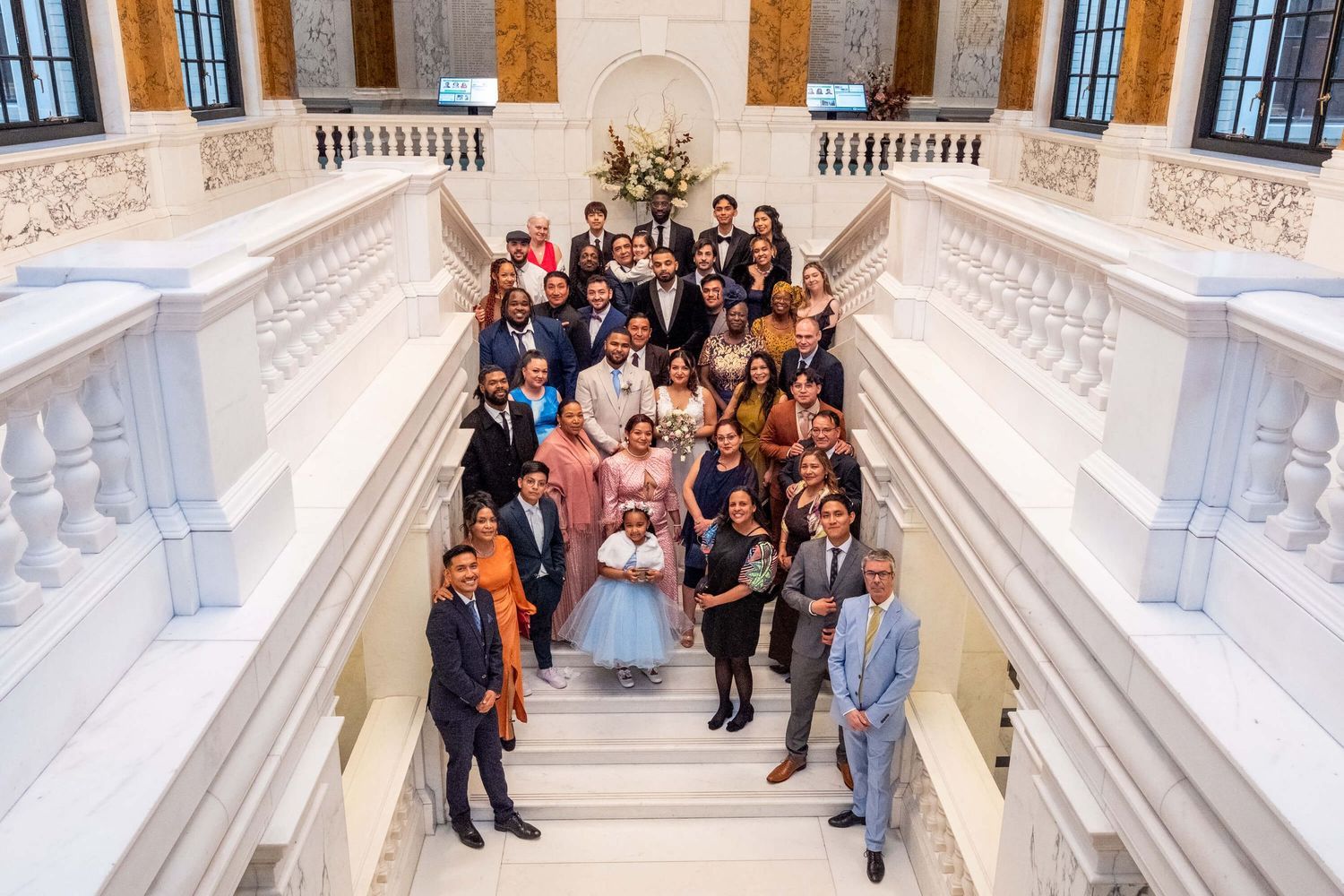 Wedding party gathers on an elegant white marble staircase in a grand historic building interior.