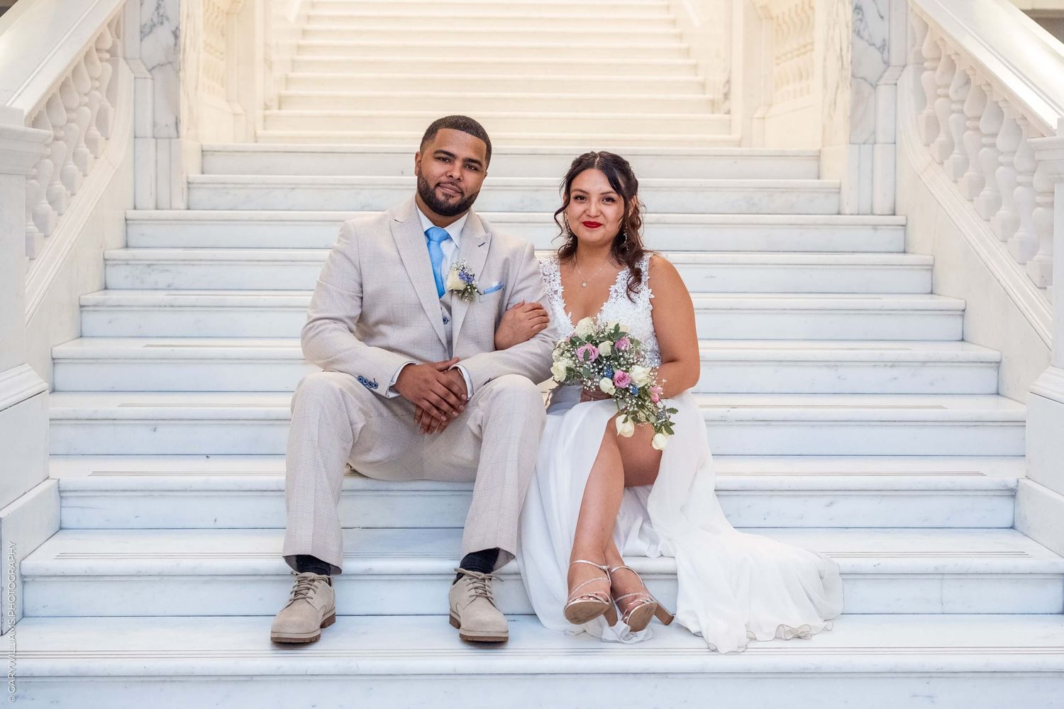 A couple sits together on white marble stairs while holding a floral bouquet at their wedding celebration.