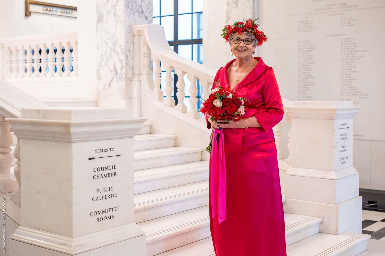 A person in a bright red dress and floral crown holds a bouquet while standing by an elegant white staircase.