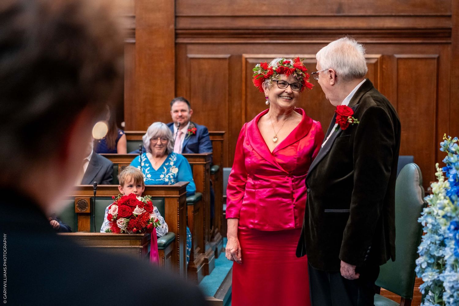 A couple stands together during a wedding ceremony in a wood-paneled room decorated with red and blue flowers.