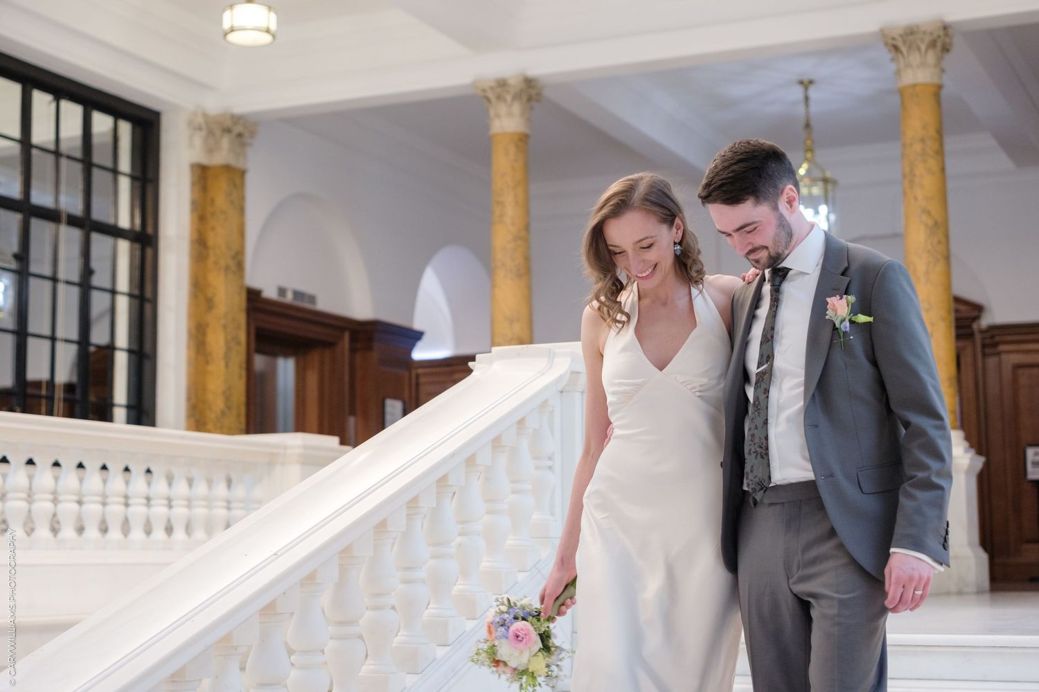 A couple descends an elegant white staircase in formal wedding attire with ornate golden pillars in the background.