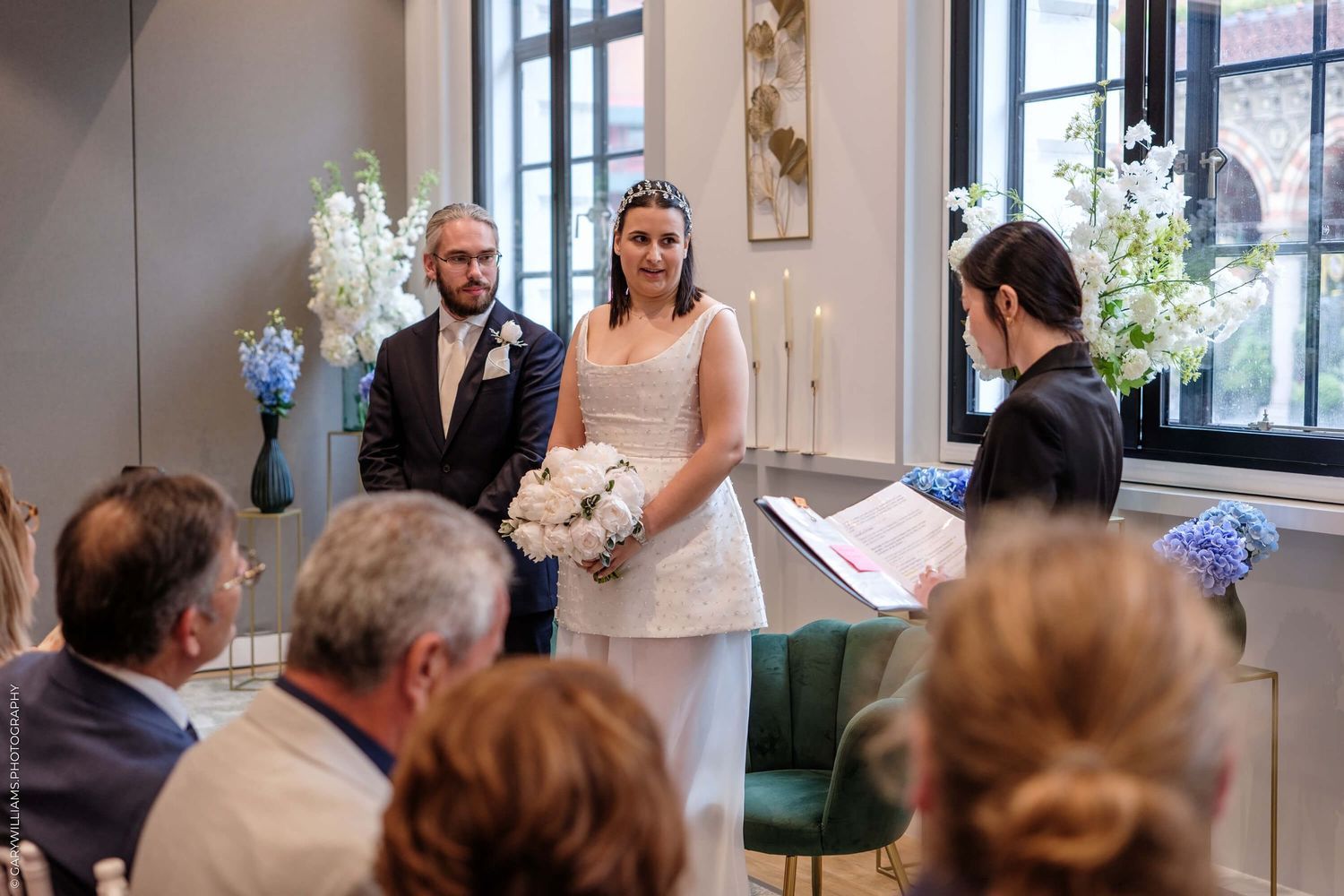 A couple stands at the front of an elegant room during an intimate wedding ceremony with blue floral arrangements.