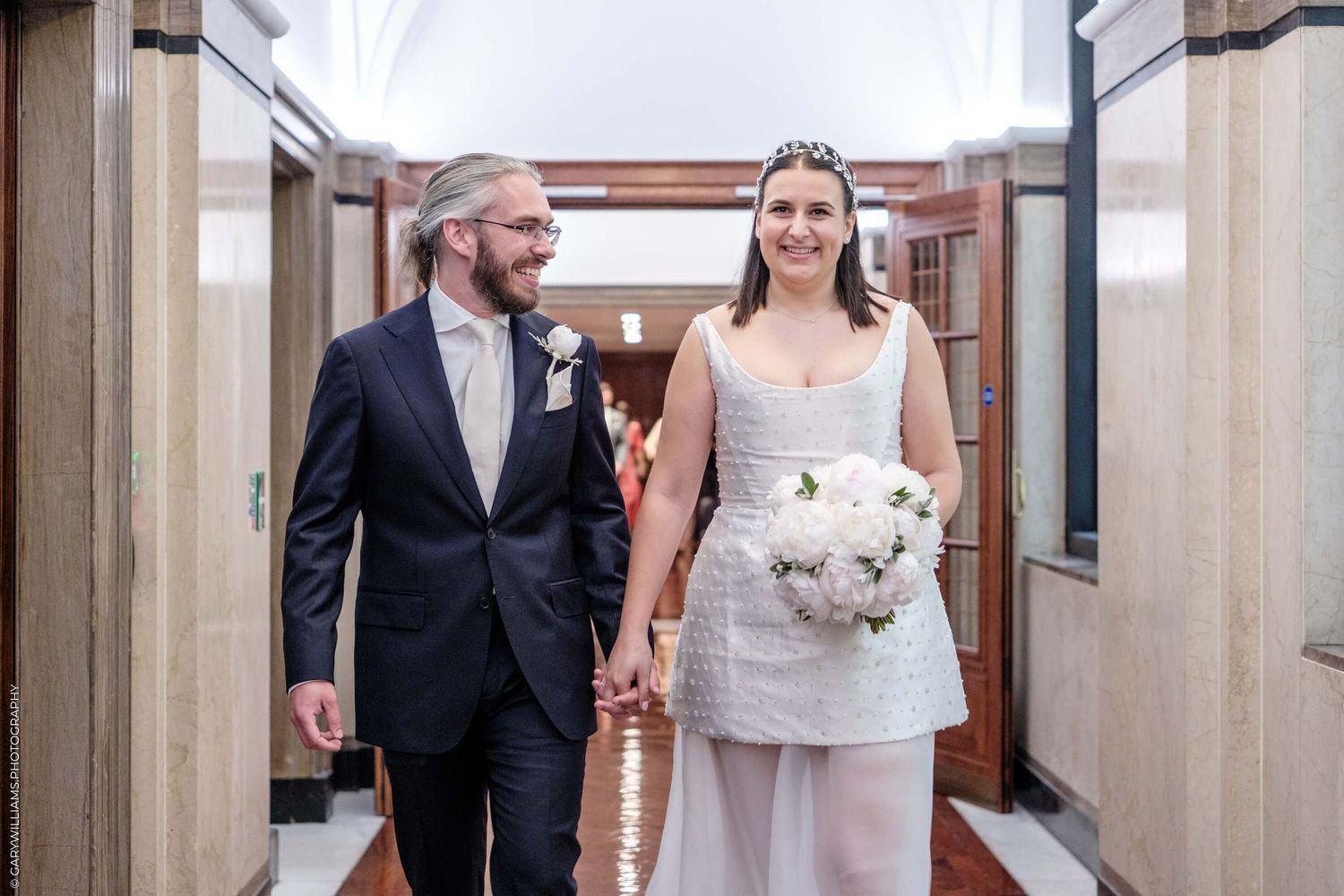 A couple walks hand-in-hand down an elegant hallway with stone columns and archways.