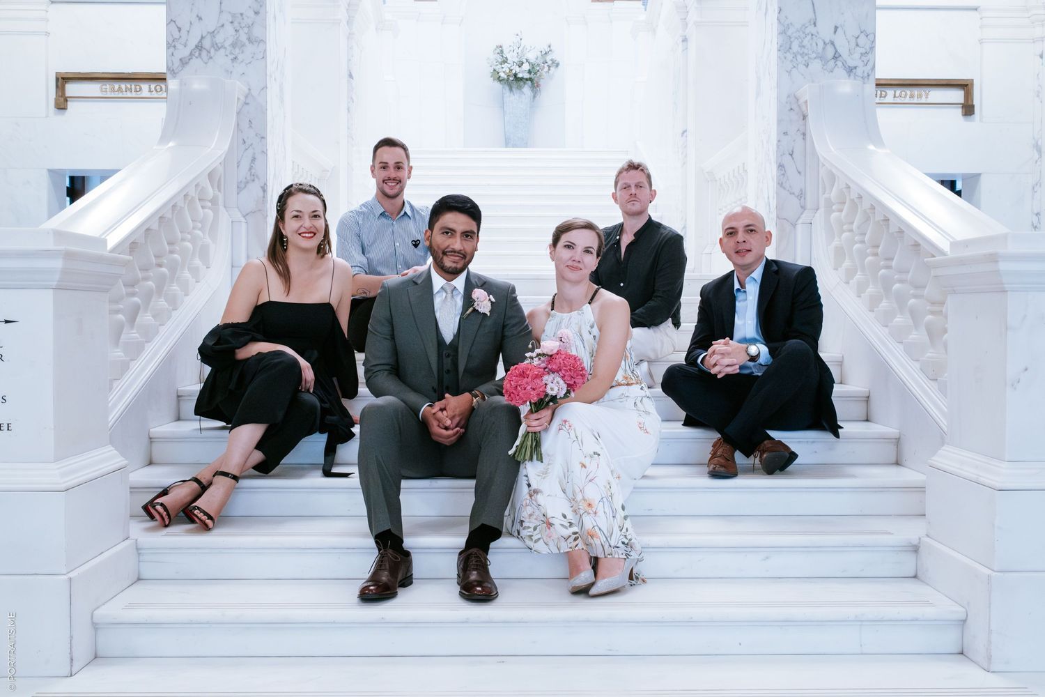 A group of six people sitting on elegant white marble steps wearing formal attire.
