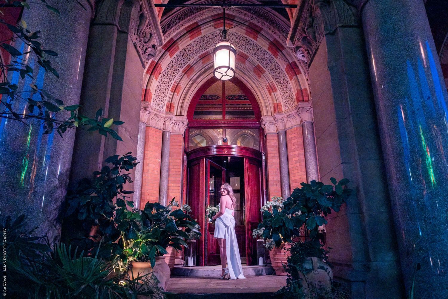 A couple stands in a grand doorway illuminated by pink and blue lighting with gothic architectural details and potted plants.