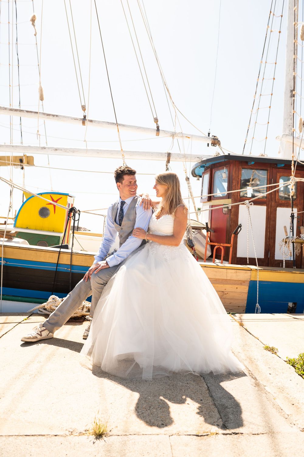 A couple in wedding attire sits together on a boat dock with a blue sailboat in the background on a sunny day.