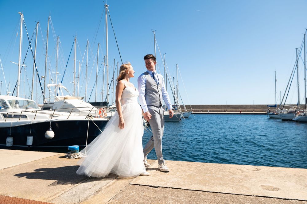 Newlyweds stand together on a sunny marina dock with sailboats and blue water in the background.
