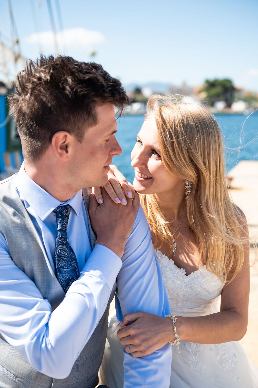 A couple shares an intimate moment on their wedding day by the waterfront, with sunny blue skies in the background.