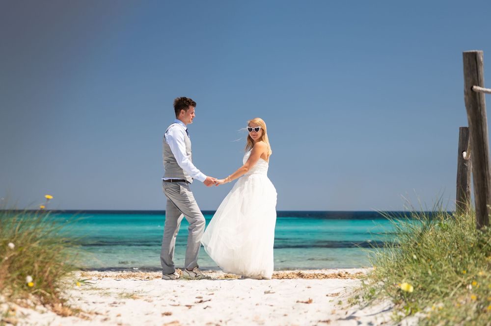 A couple in wedding attire stands together on a white sandy beach with turquoise ocean waters and beach grass in view.