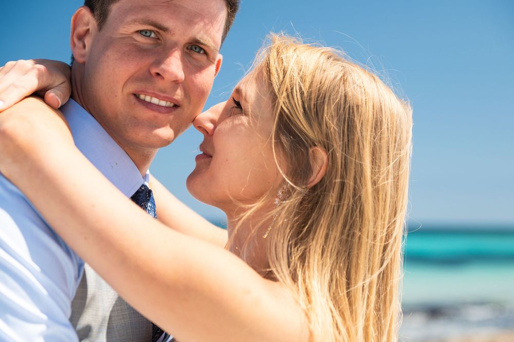 A couple embraces lovingly on a sunny beach with turquoise ocean waters in the background.