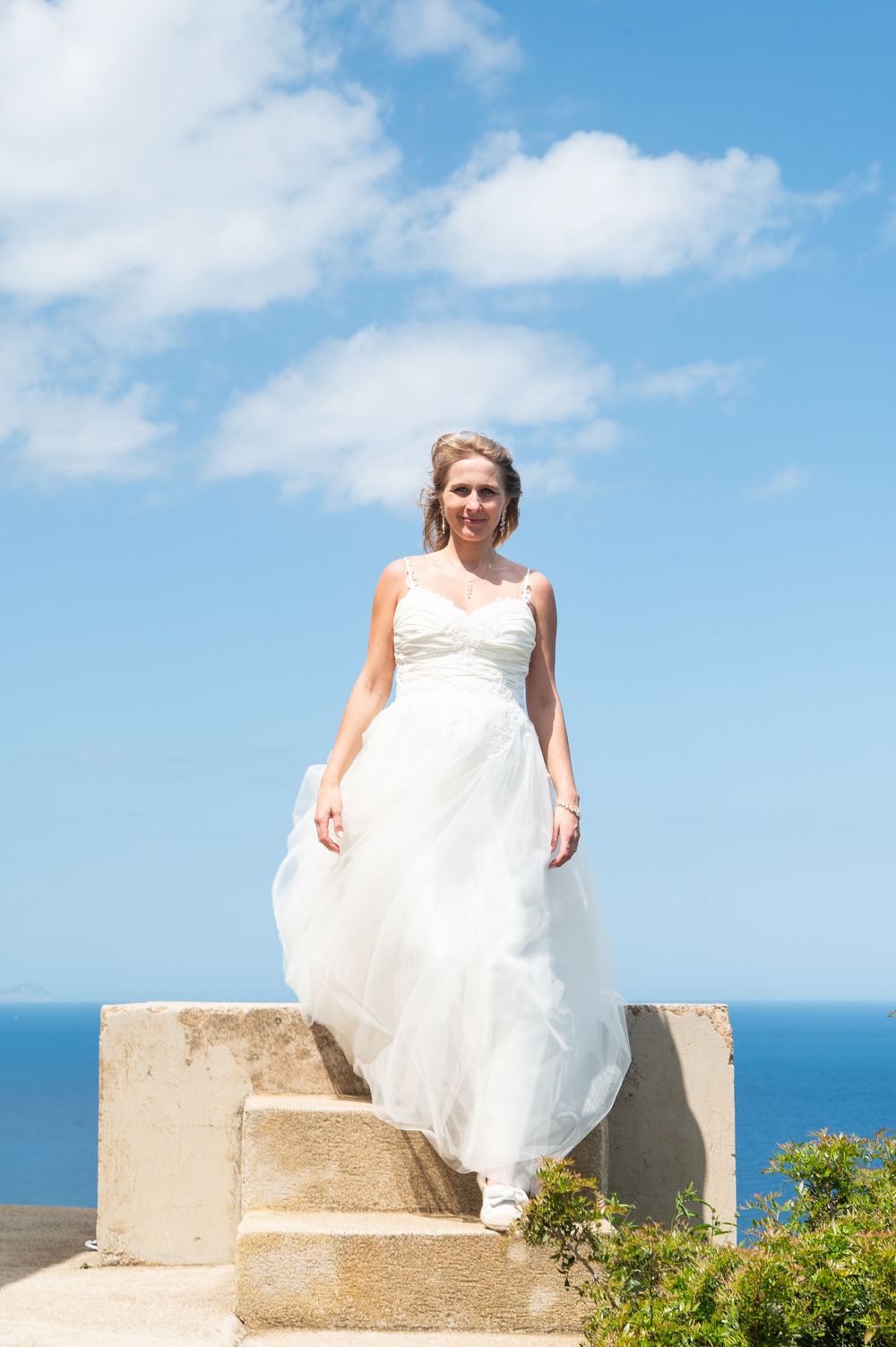 A bride in a flowing white wedding dress stands atop stone steps overlooking a bright blue ocean vista.
