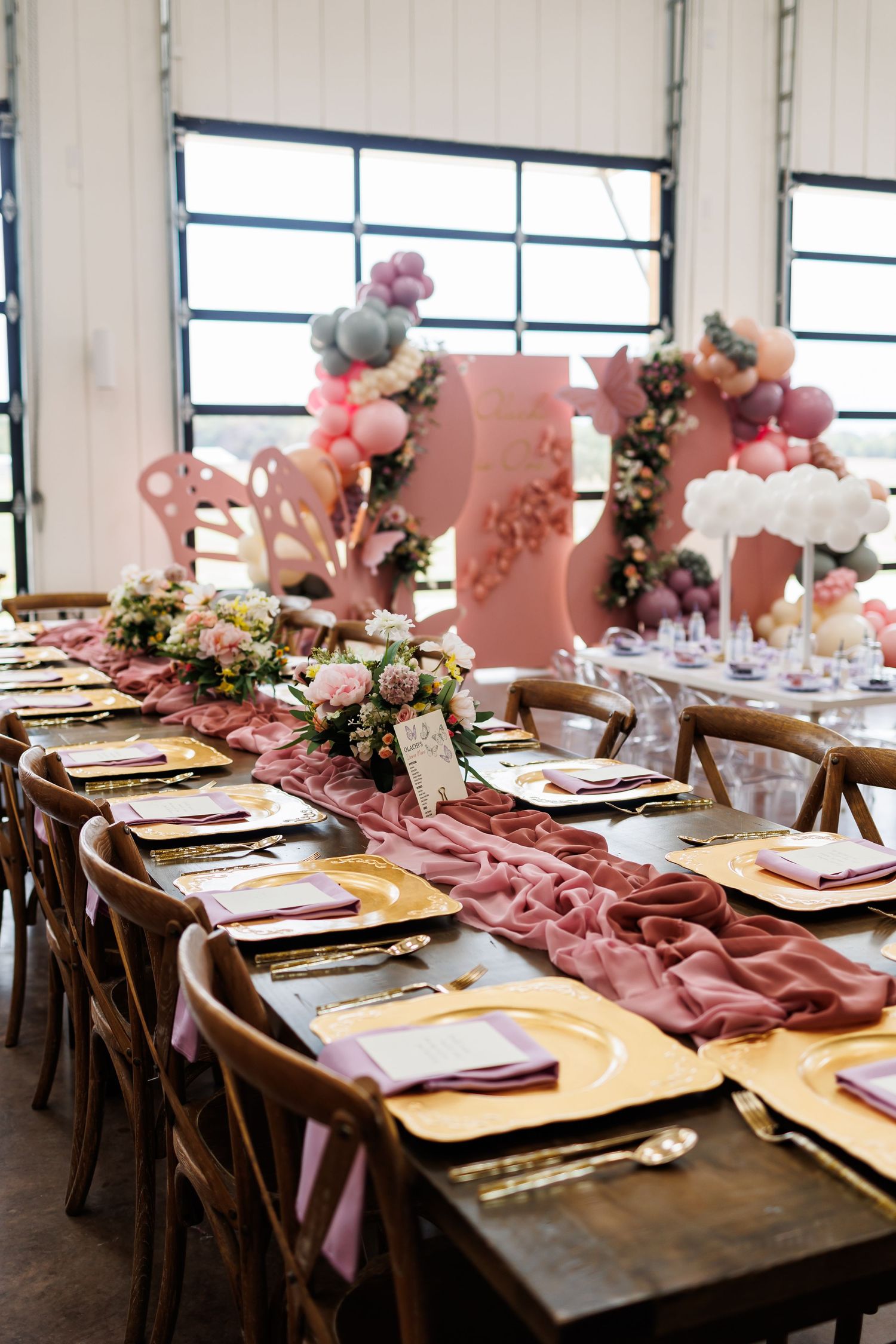 Pink and purple butterfly-themed tablescape with pink fabric runner, gold chargers, and balloon arch decorations at Bella Cavalli
