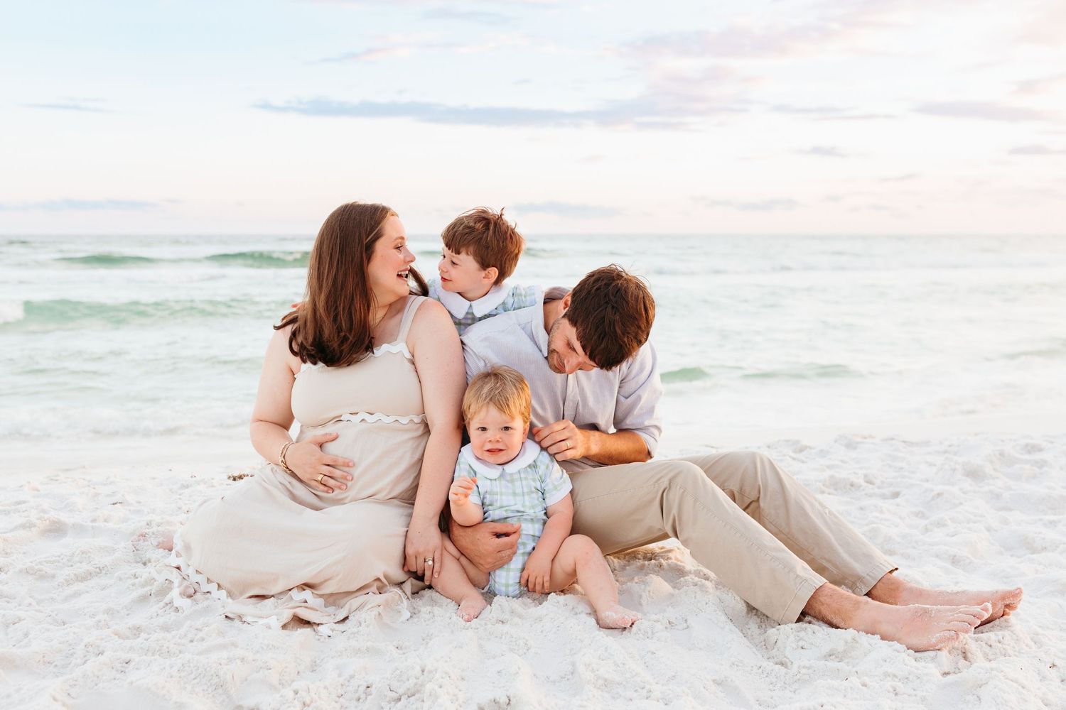 A family sits together on white sand beach at sunset wearing neutral colored clothing.
