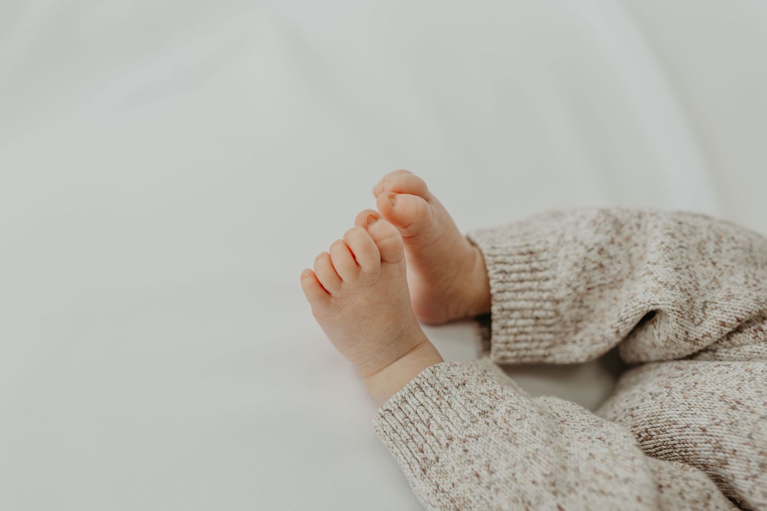 Tiny baby foot against white bedding with cozy beige knit sweater sleeve in view. London and beckenham photographer.