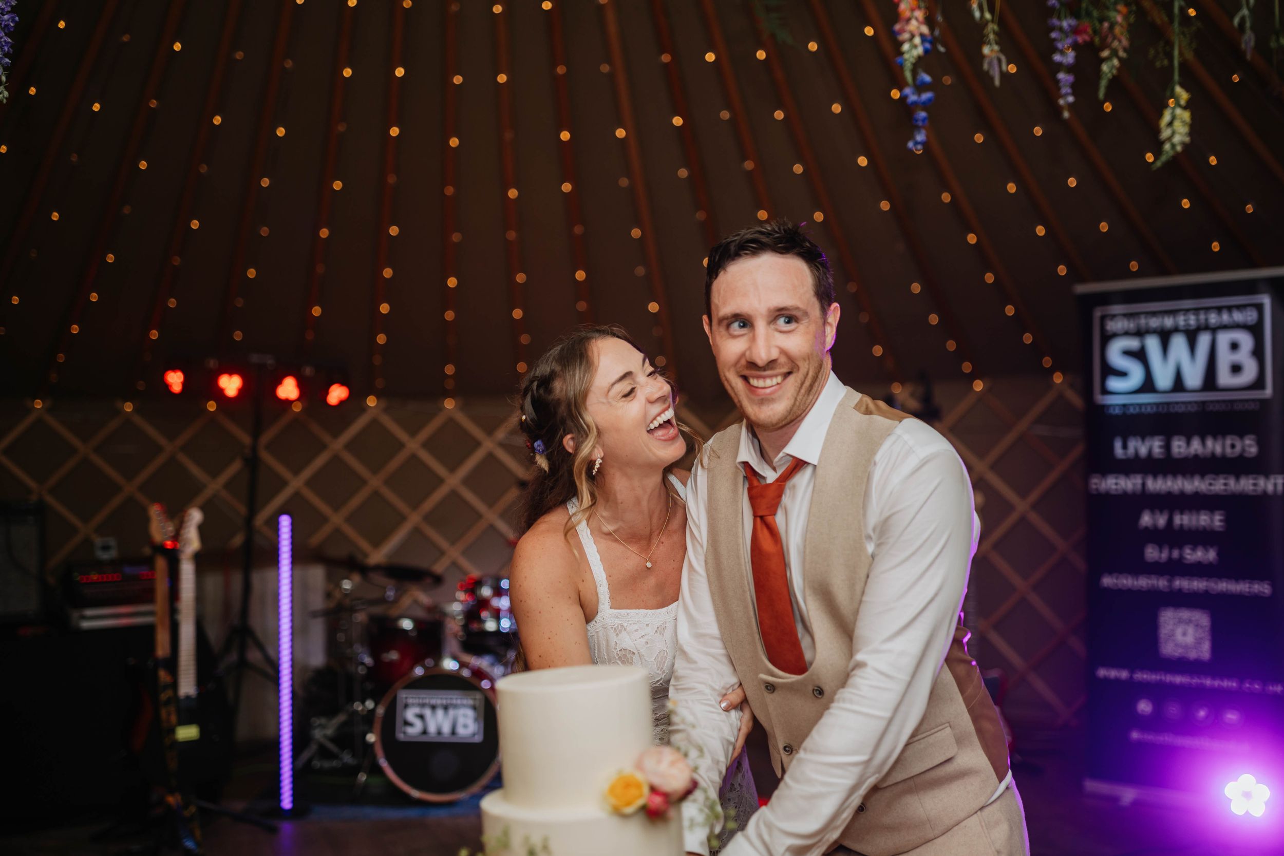 Joyful couple shares a candid dance moment at their wedding reception with twinkling lights and SWB signage in background.