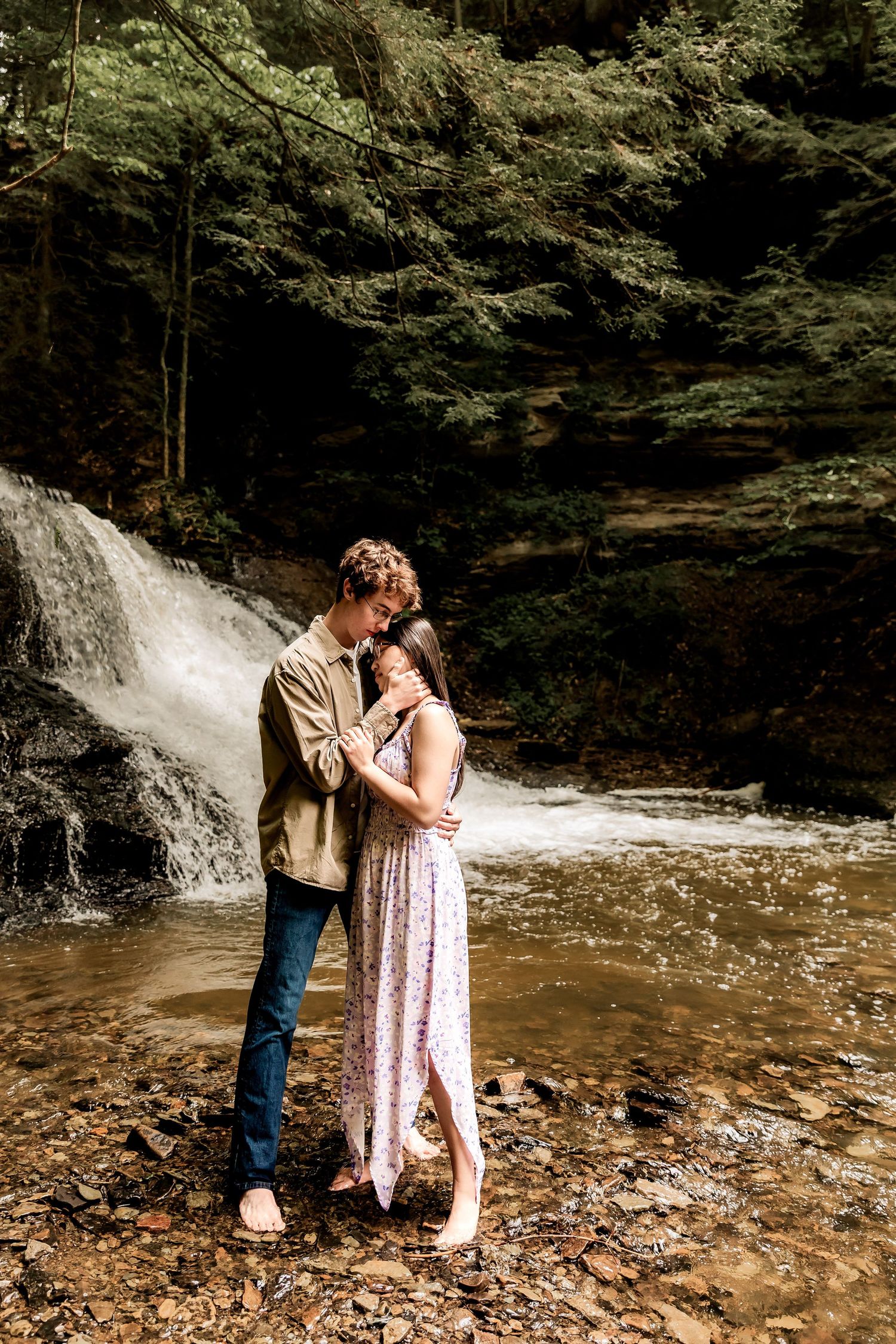 A romantic couple embraces near a cascading waterfall surrounded by lush green forest.