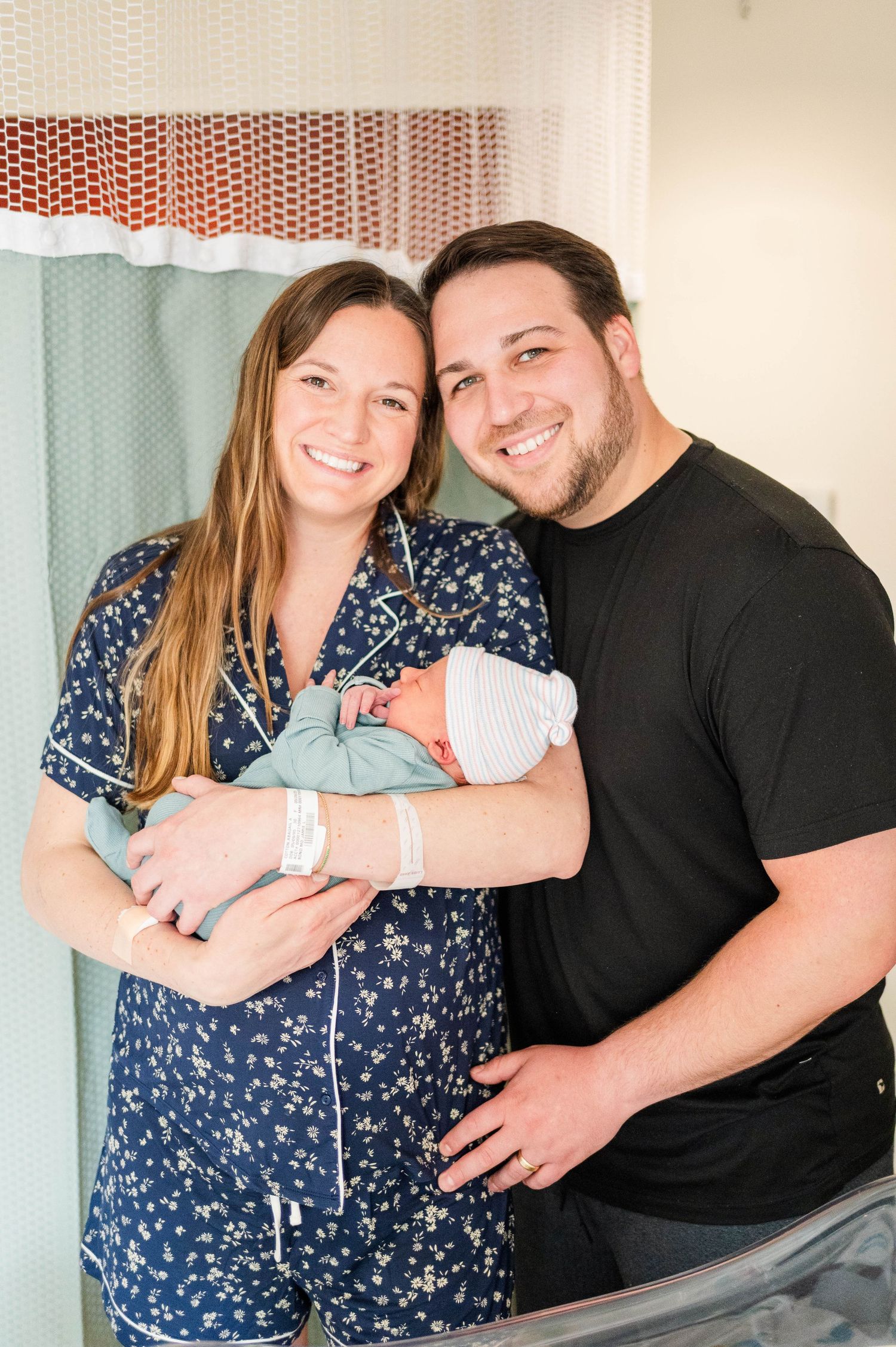 A couple in a hospital room holds their newborn baby, wearing a blue and black floral dress and black t-shirt.