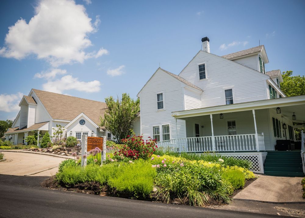 A panoramic view of white farmhouse-style buildings nestled along a hillside with lush green landscaping under blue skies.