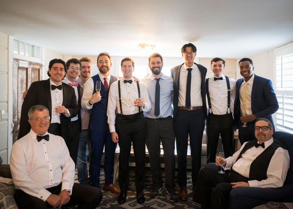 A group in formal attire including bow ties and suspenders pose together in a bright indoor setting.