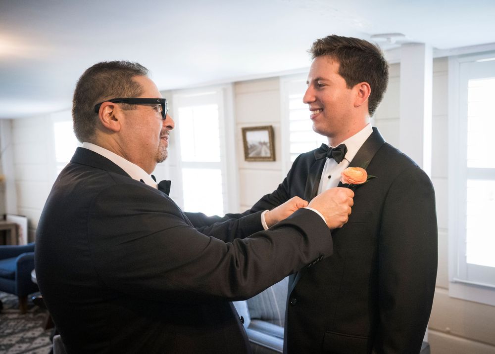 A person helps adjust another person's boutonniere and tie before a formal event in a bright indoor setting.