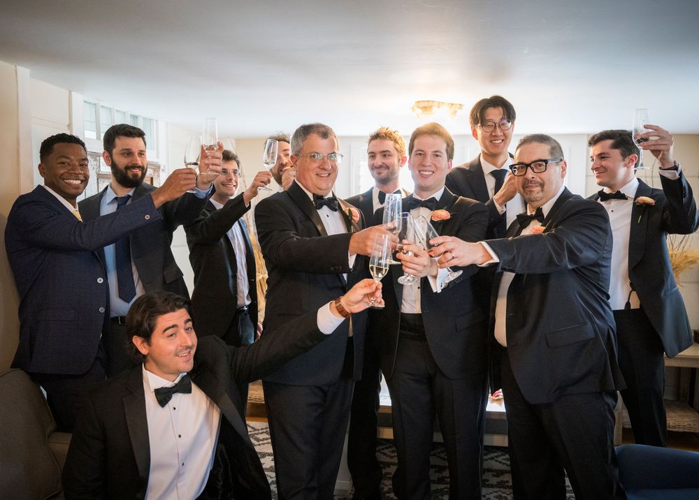 Wedding groomsmen in black tuxedos toast and celebrate during a candid moment at reception.