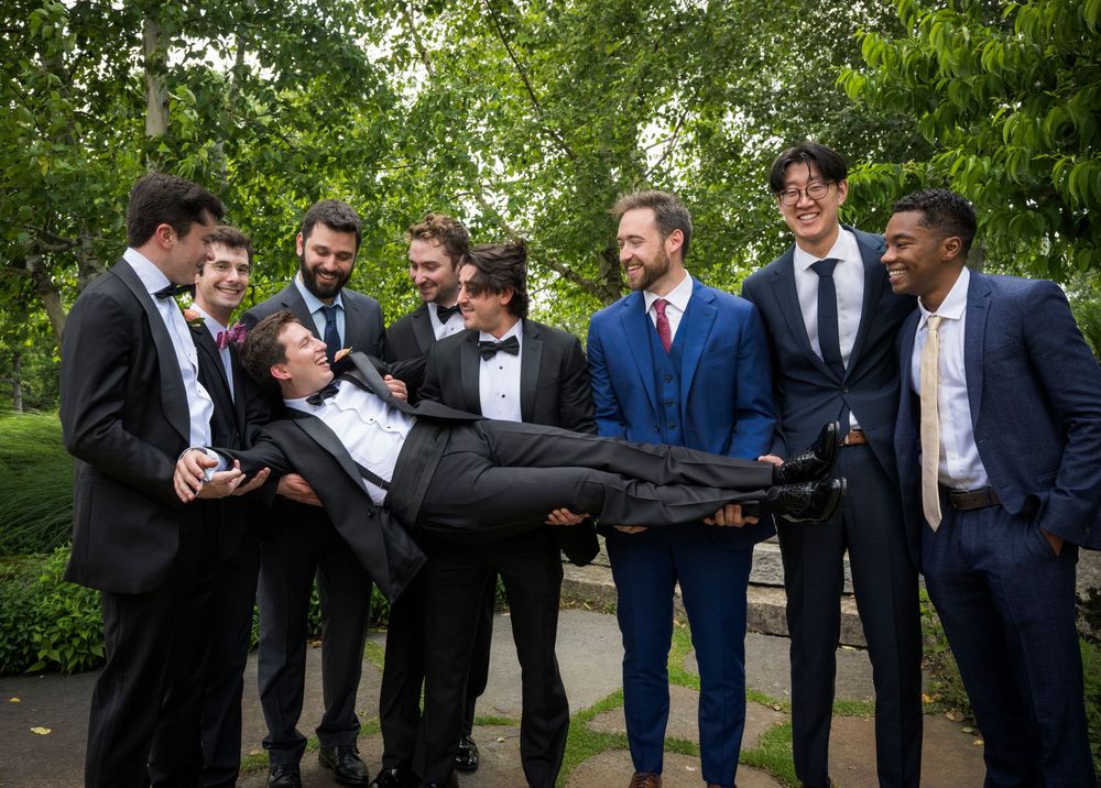 Groomsmen in dark suits stand together outdoors sharing drinks and smiles at an elegant wedding celebration.