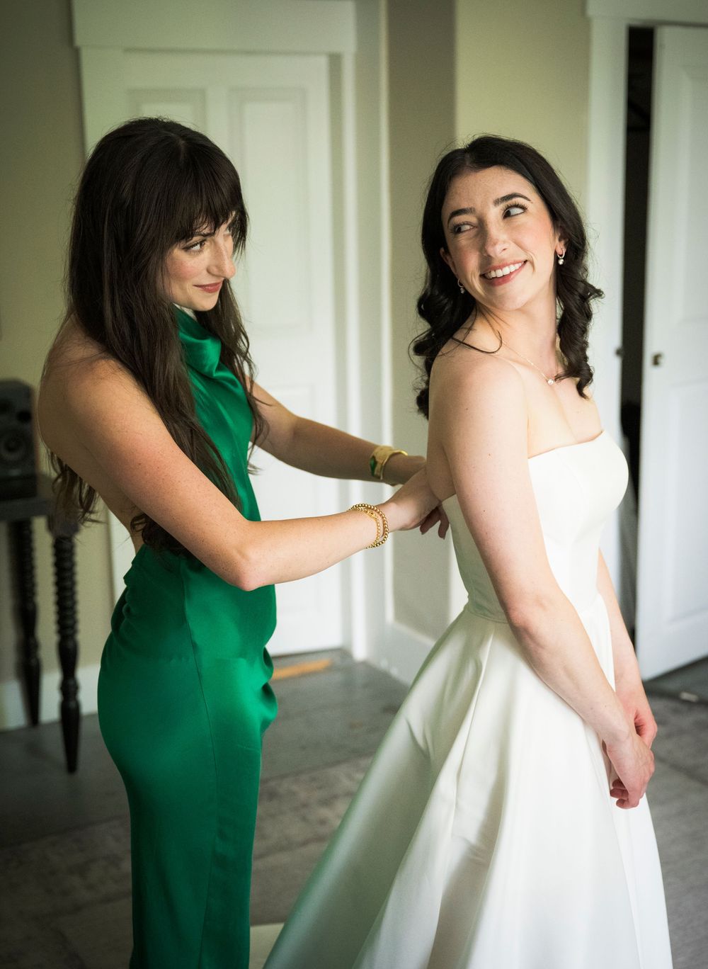 Two figures in green and white dresses pose playfully in an elegant indoor setting next to white doors and a staircase.