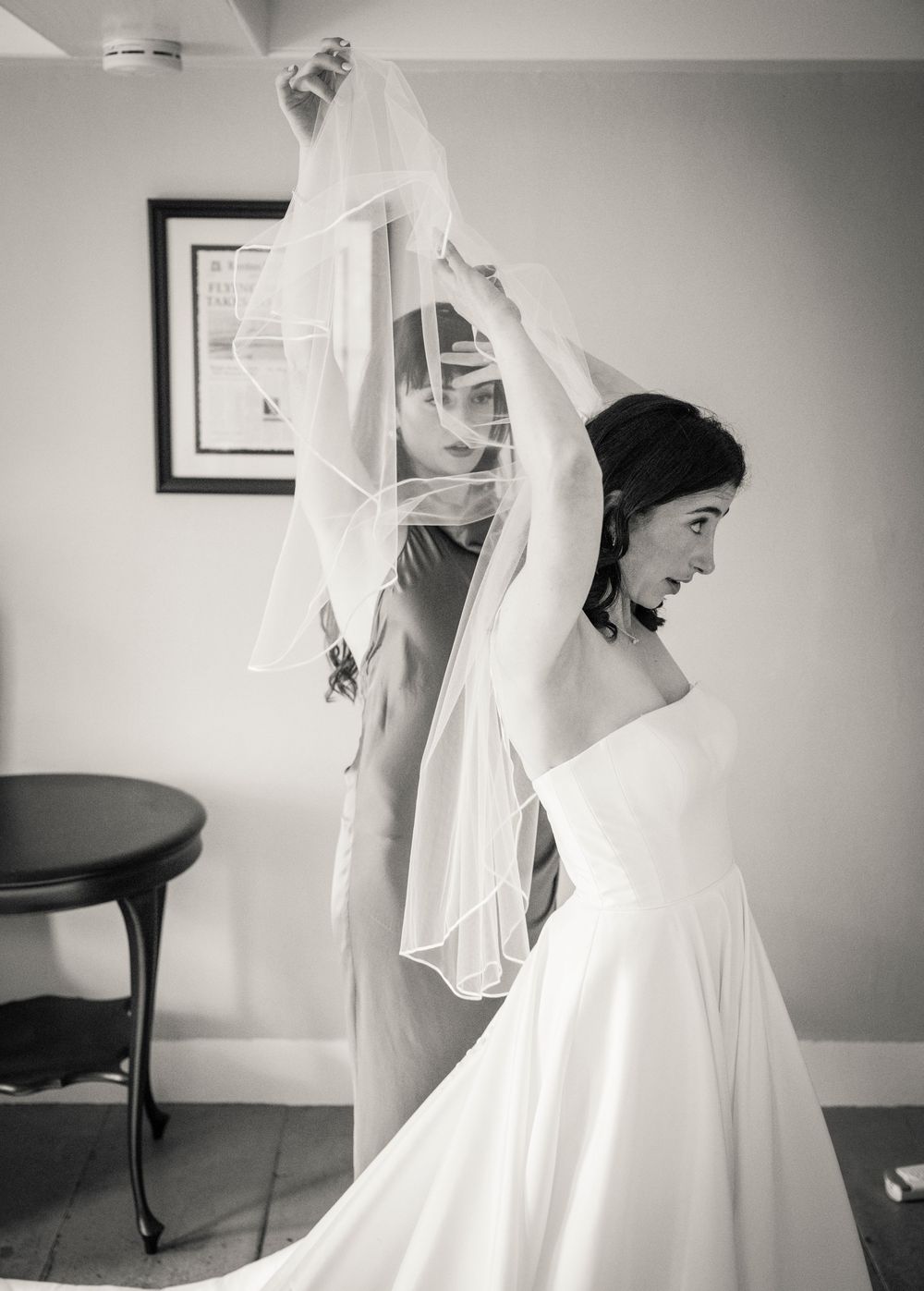 A graceful bride in a flowing white gown and veil stands in soft lighting during the wedding preparation.