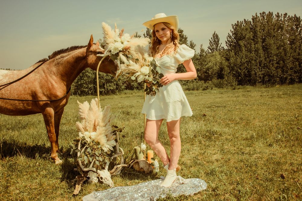 A person in a white dress stands in a meadow next to a brown horse with floral arrangements in vintage-style lighting.