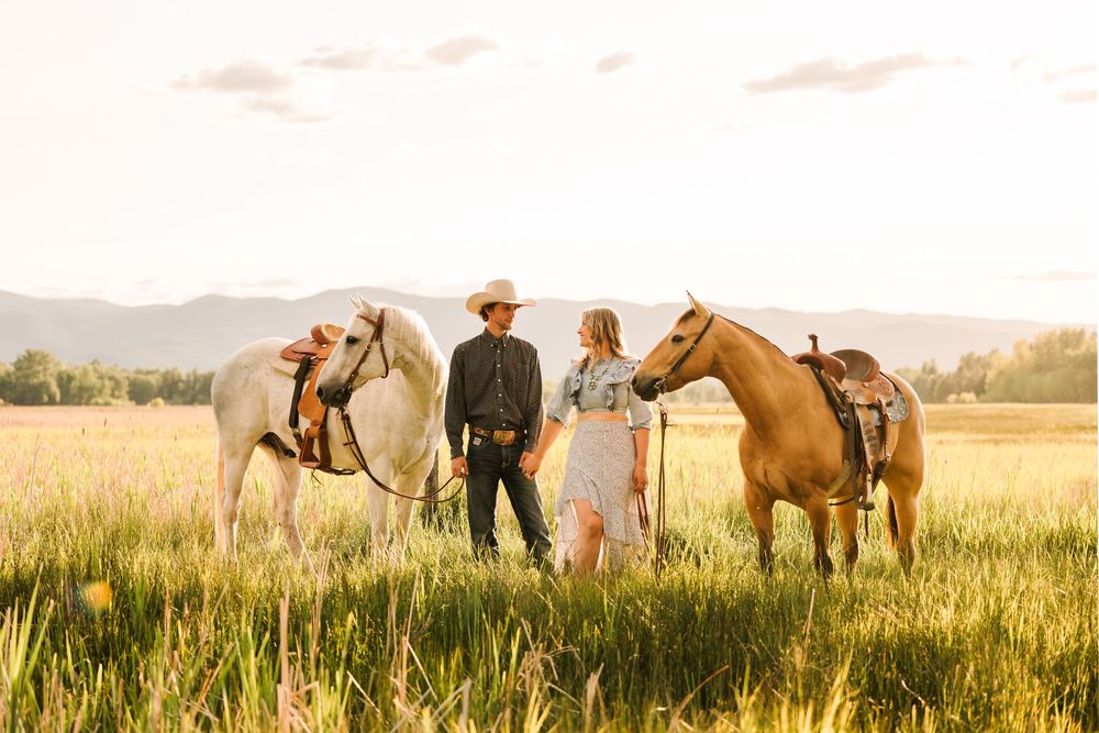 Couple stands with two saddled horses in a golden wheat field at sunset with mountains in the background.