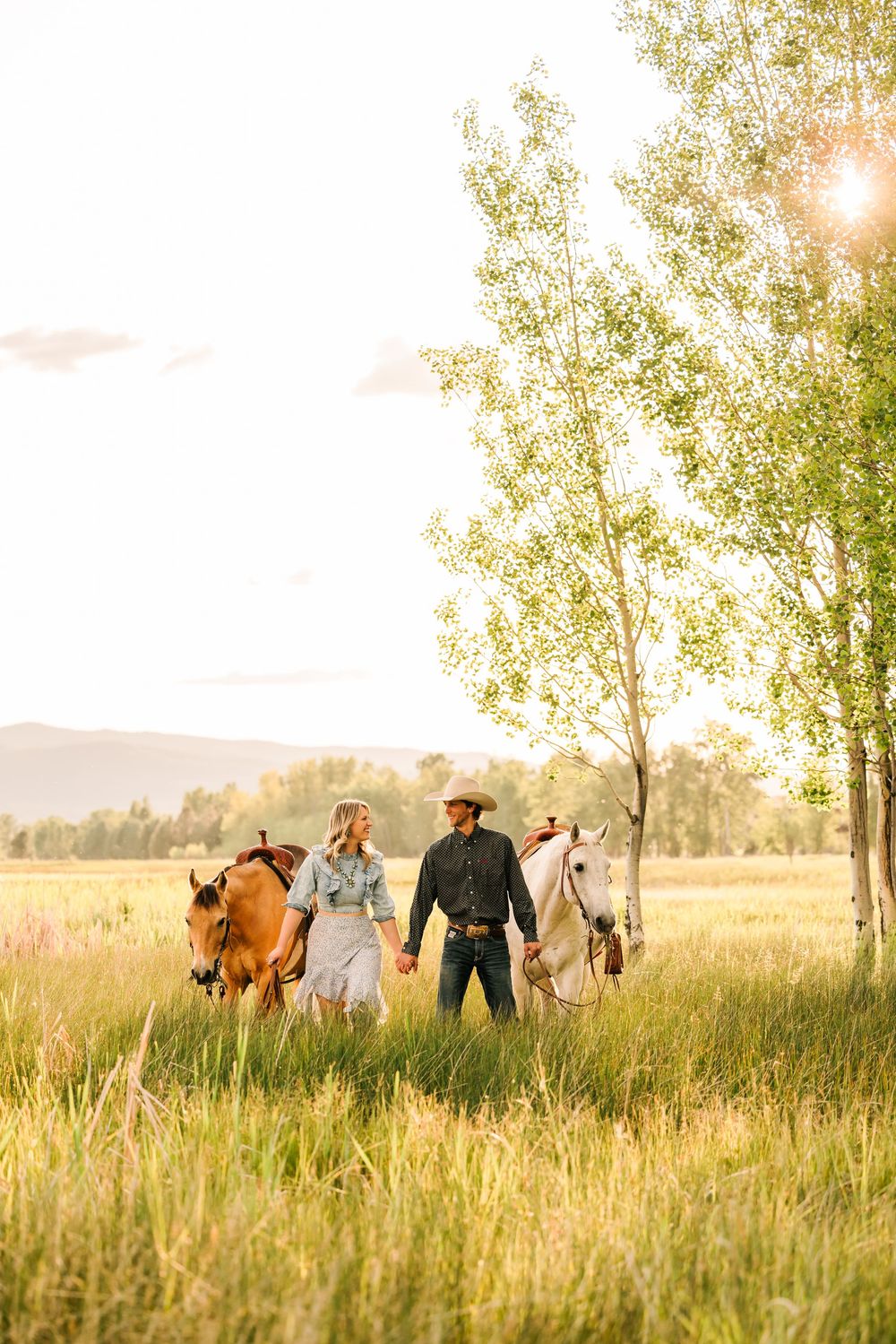 Two riders in western attire lead horses through a golden meadow at sunset with tall trees in the background.
