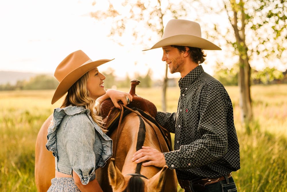 Two people in cowboy hats share a romantic moment on horseback in a sunlit field at golden hour.
