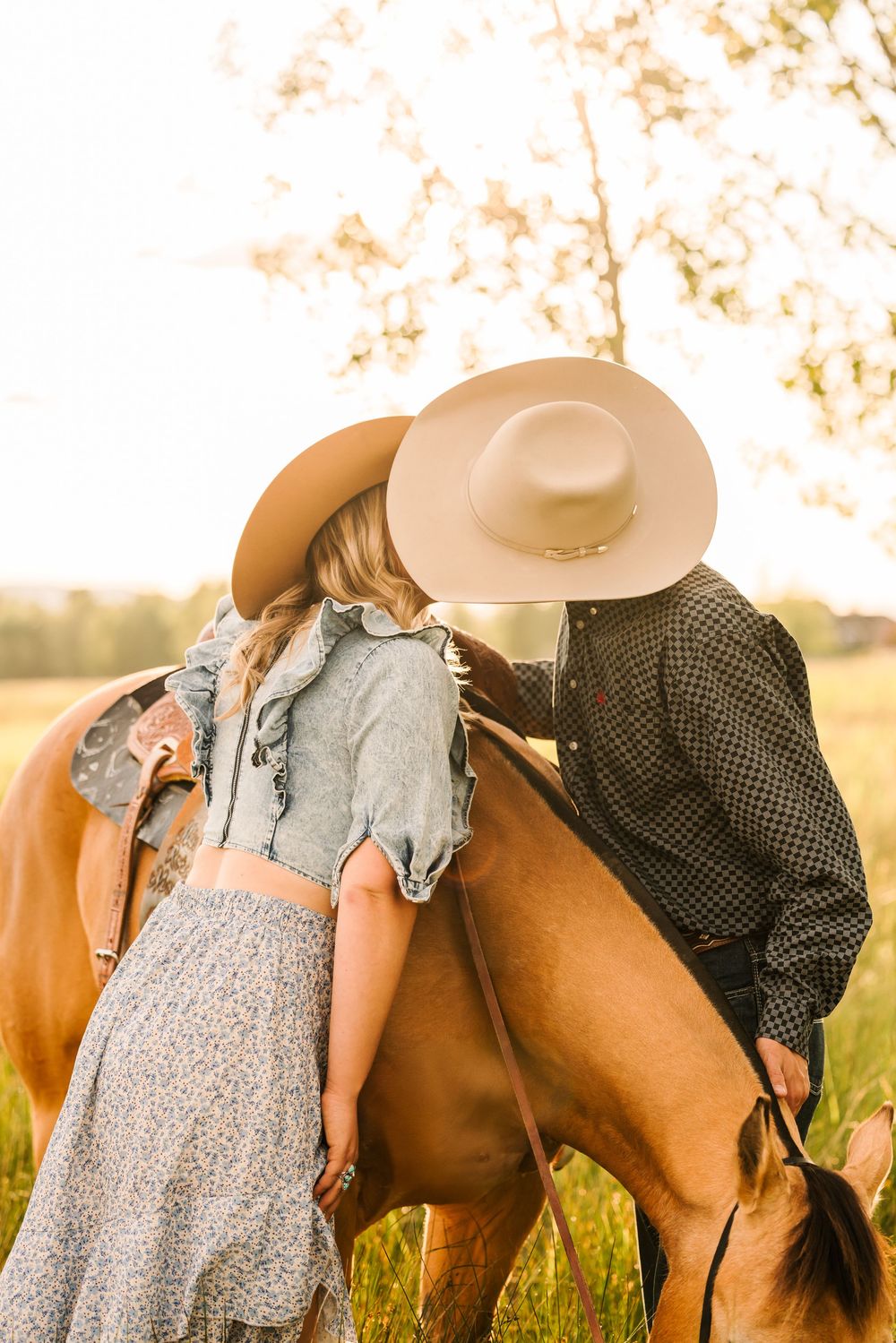 A couple in western attire poses with a brown horse during a golden sunset in a grassy field.