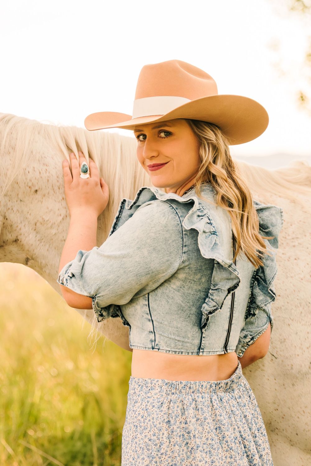 Woman in denim jacket and floral dress poses in golden sunlight wearing brown cowboy hat against rustic wall.