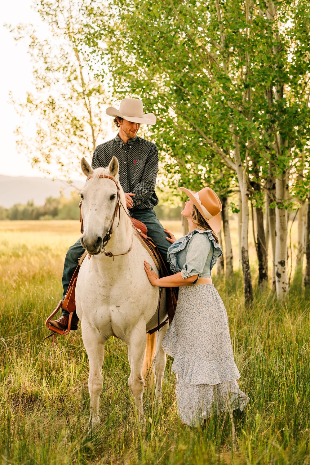 An intimate countryside portrait with horses, cowboy hats, and a flowing dress among birch trees.