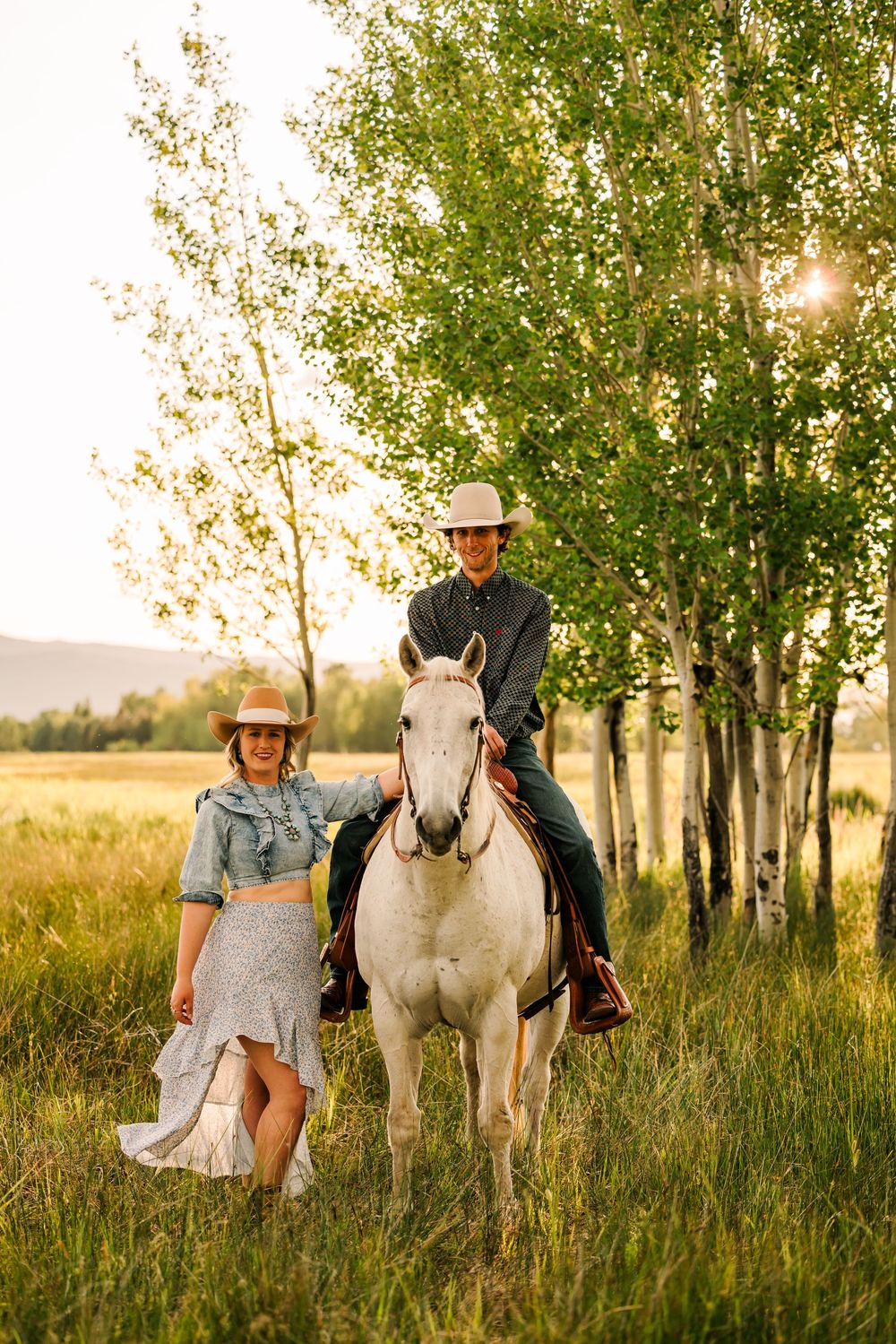 Couple in western attire poses with a white horse in a meadow at sunset with aspen trees in the background.