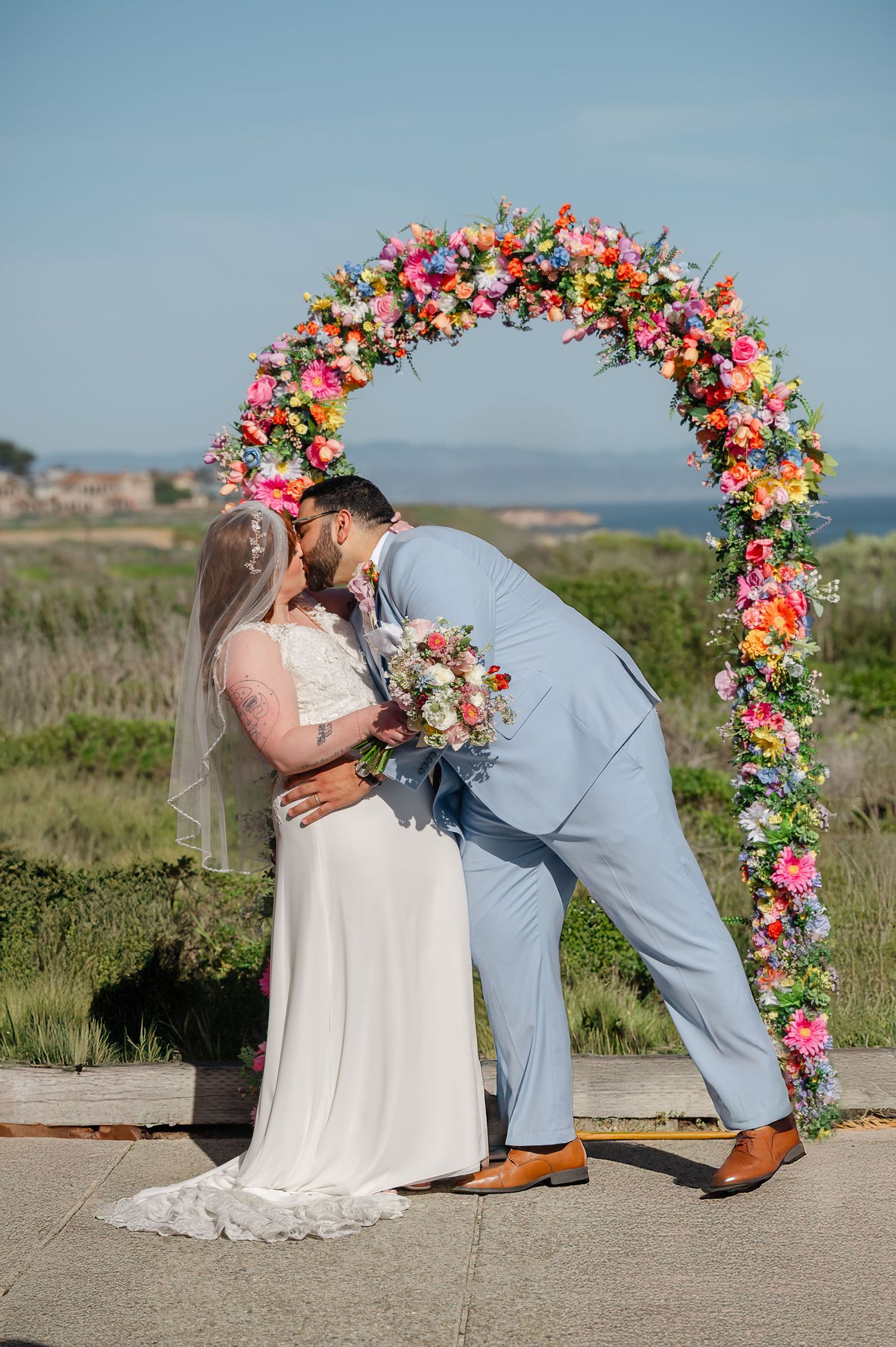 A couple in wedding attire share romantic moments in a scenic outdoor setting with vibrant floral arch decorations.