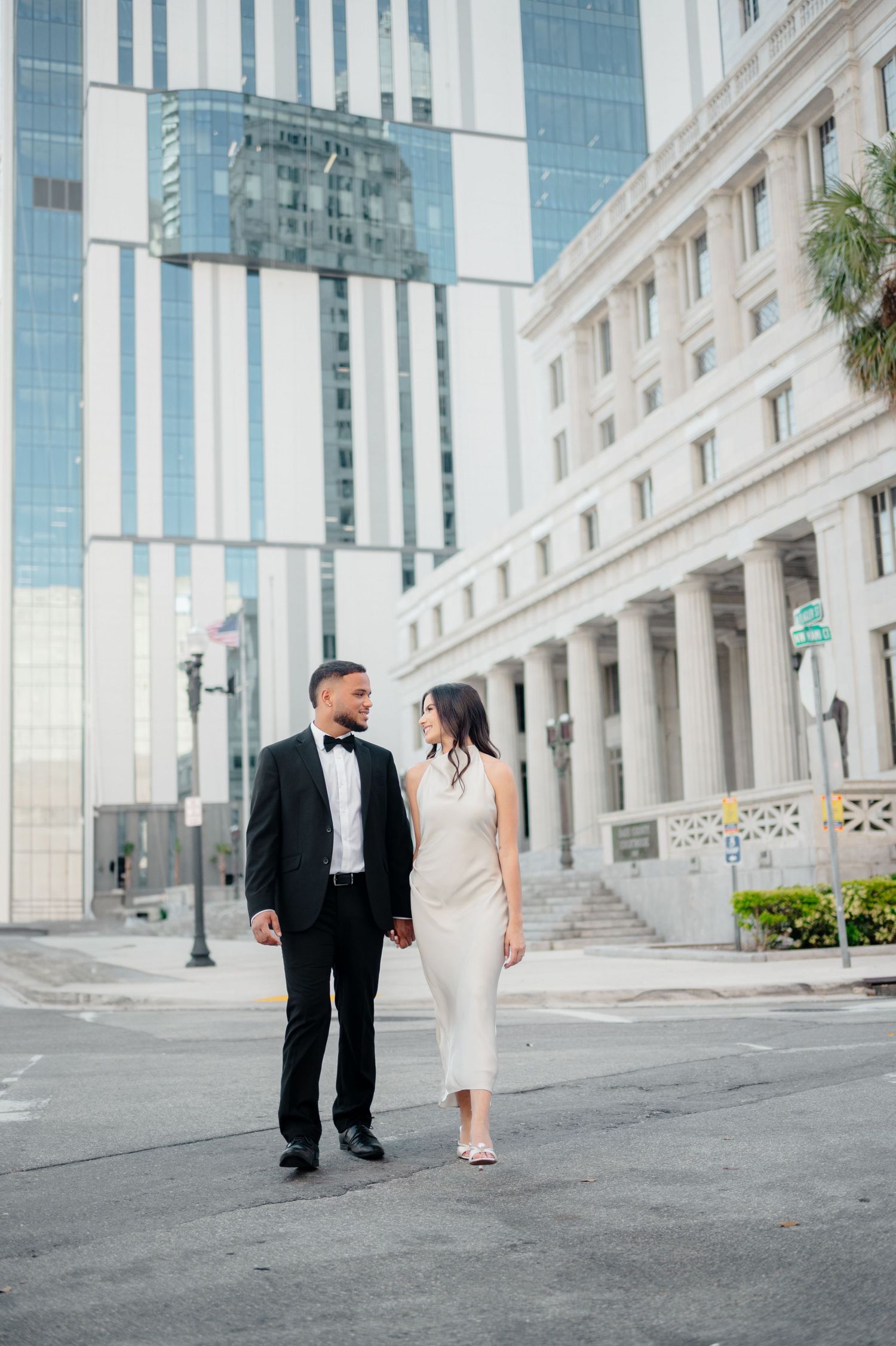 A formal couple in black and white attire poses elegantly in front of a modern architectural building downtown.