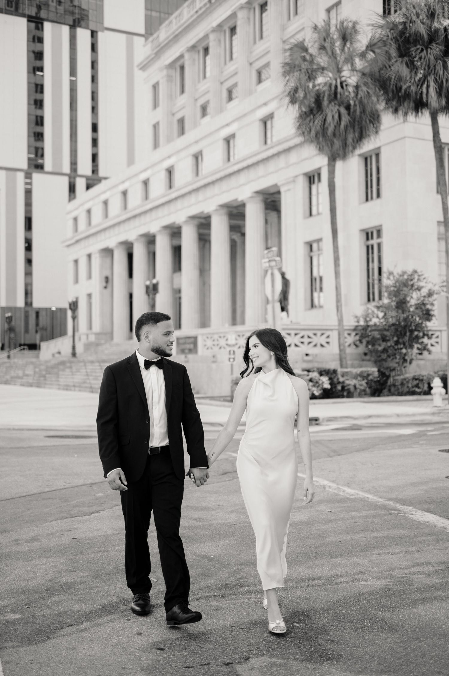 Black and white photo of a couple in formal attire walking hand in hand outside a grand neoclassical building with columns.