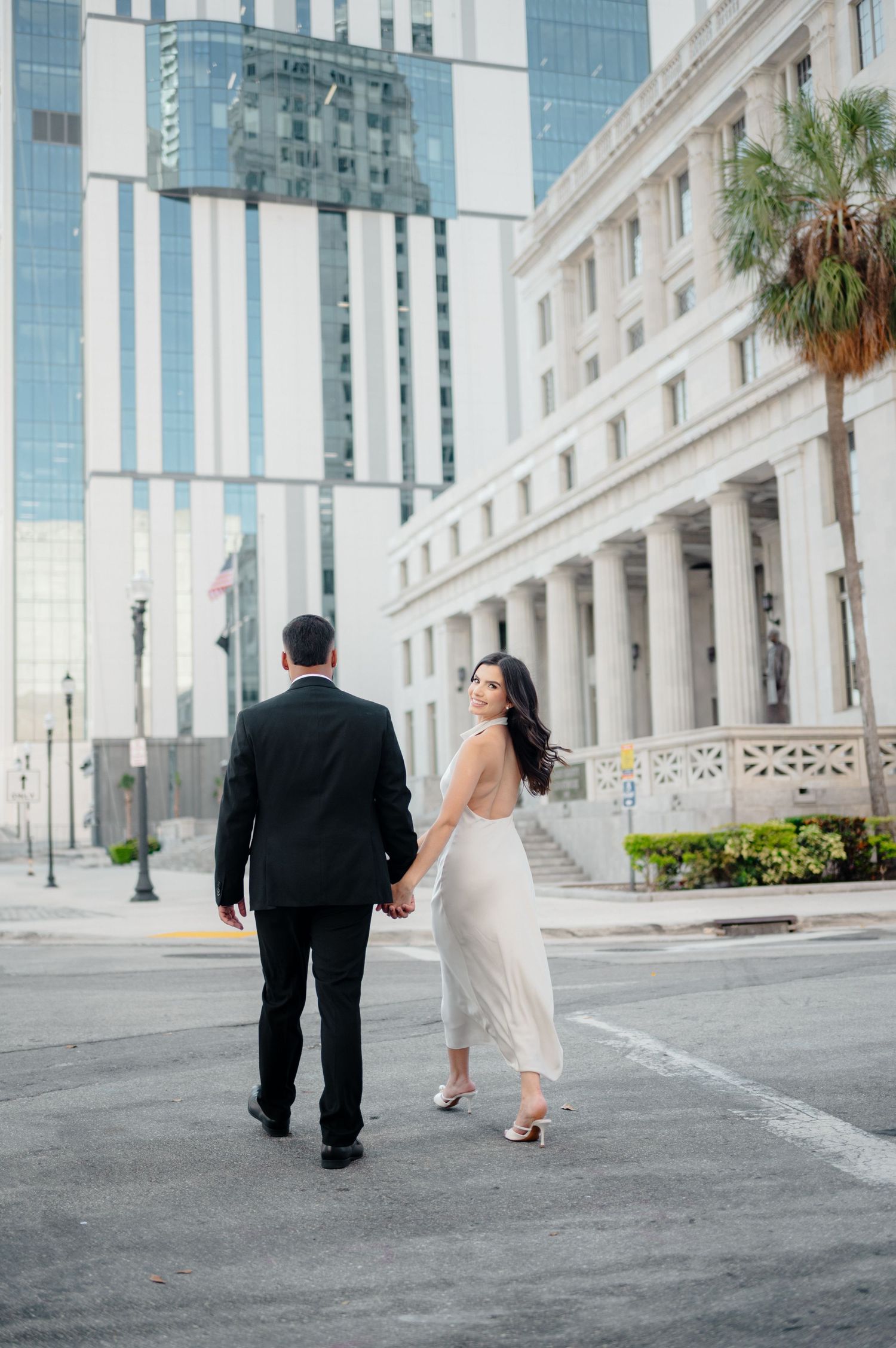 A couple in wedding attire walk together on a city street with historic architecture and modern buildings in the background.