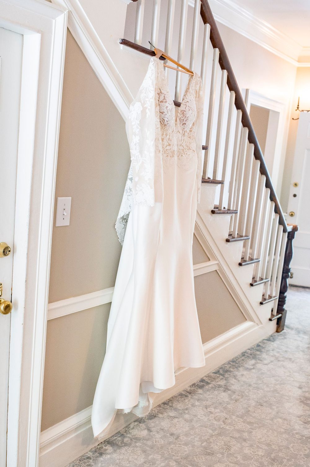 A white lace wedding dress hangs elegantly on a staircase railing with wooden banister in a neutral-toned hallway. Primrose Cottage Wedding