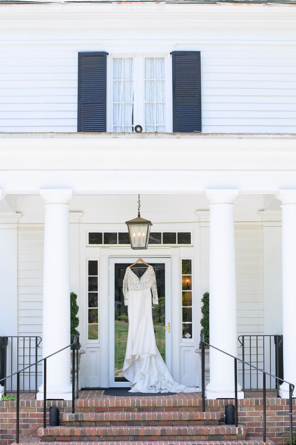 An elegant white wedding dress hangs on display in the grand entrance of a classic Southern colonial style home. Primrose Cottage Wedding