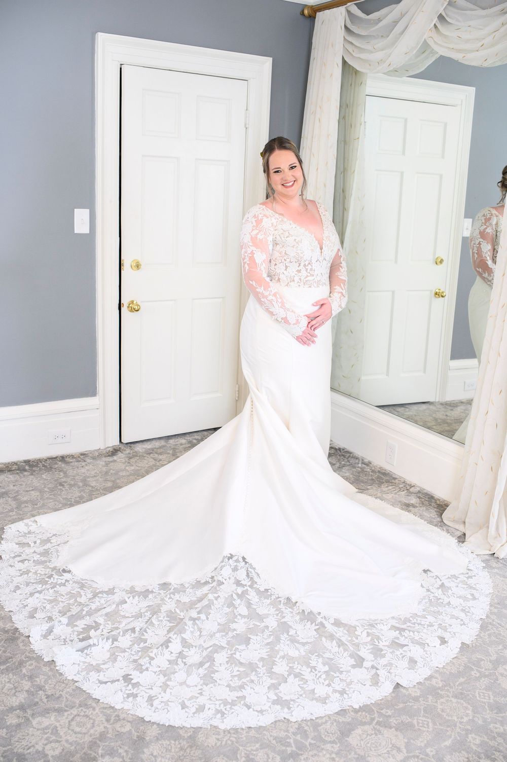 A bridal gown with long lace sleeves and dramatic cathedral train photographed in fitting room with gray walls and mirror. Primrose Cottage