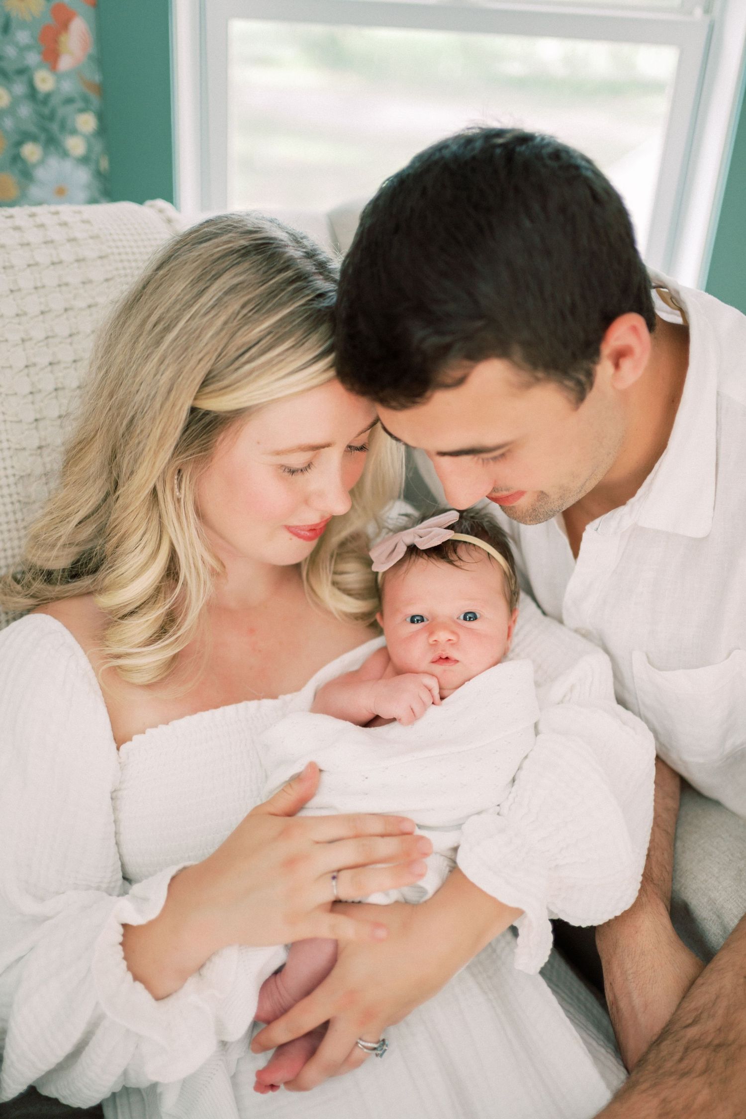 An adorable newborn baby wearing a bow headband is cradled in soft white blankets while being lovingly embraced.