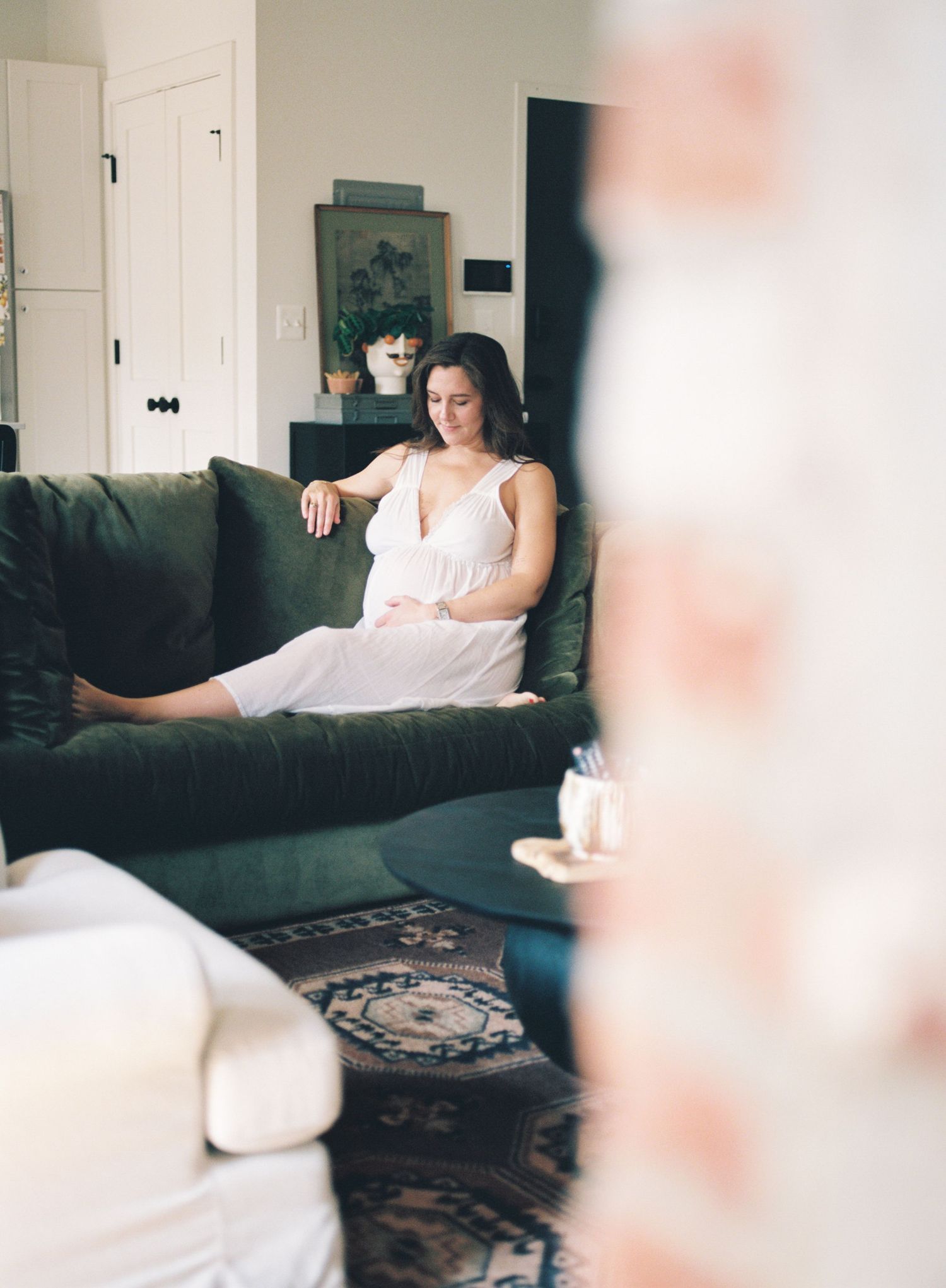 Film photograph of someone in a white dress lounging on a dark green couch in a bright, airy living room.