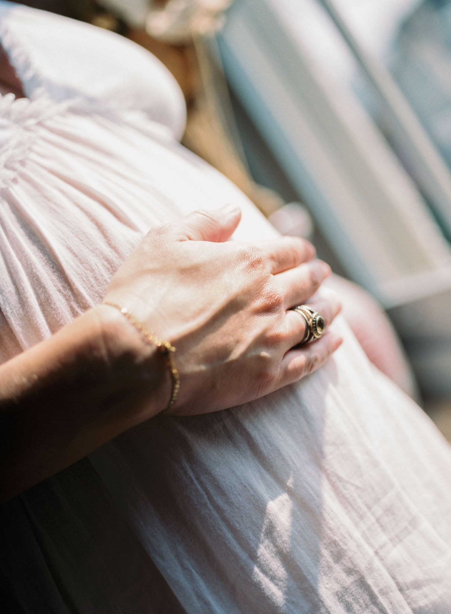 A delicate feminine hand with a gold bracelet and rings rests on soft white fabric with gentle sunlight filtering through.