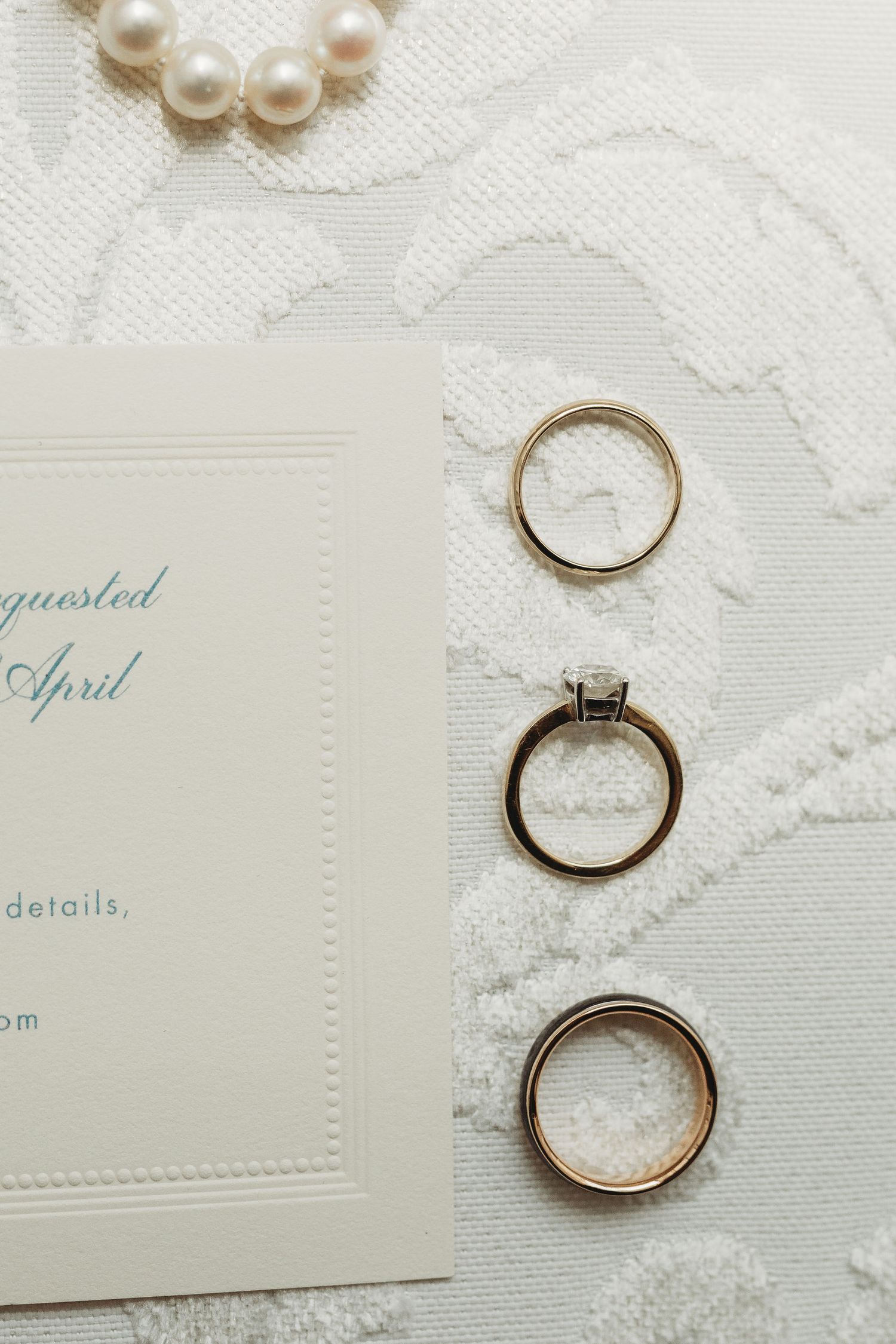 Wedding rings and pearls arranged on white lace fabric next to an elegant invitation card with April details.