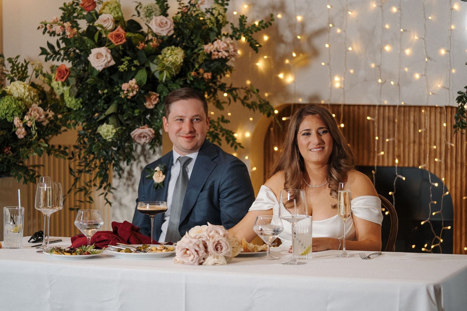 Two guests speak at a microphone during a wedding reception with floral decorations and twinkle lights in the background.