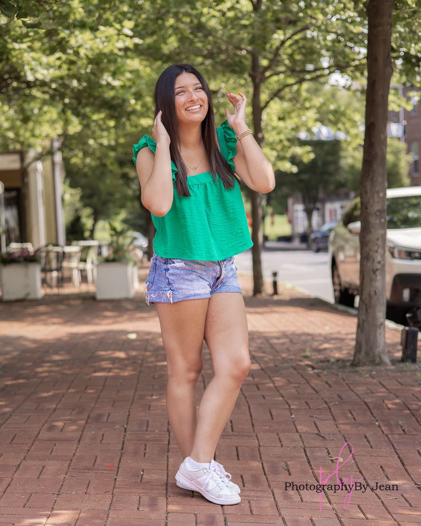 Happy person poses casually on a brick sidewalk wearing a bright green top, jean shorts and white sneakers on a sunny day.