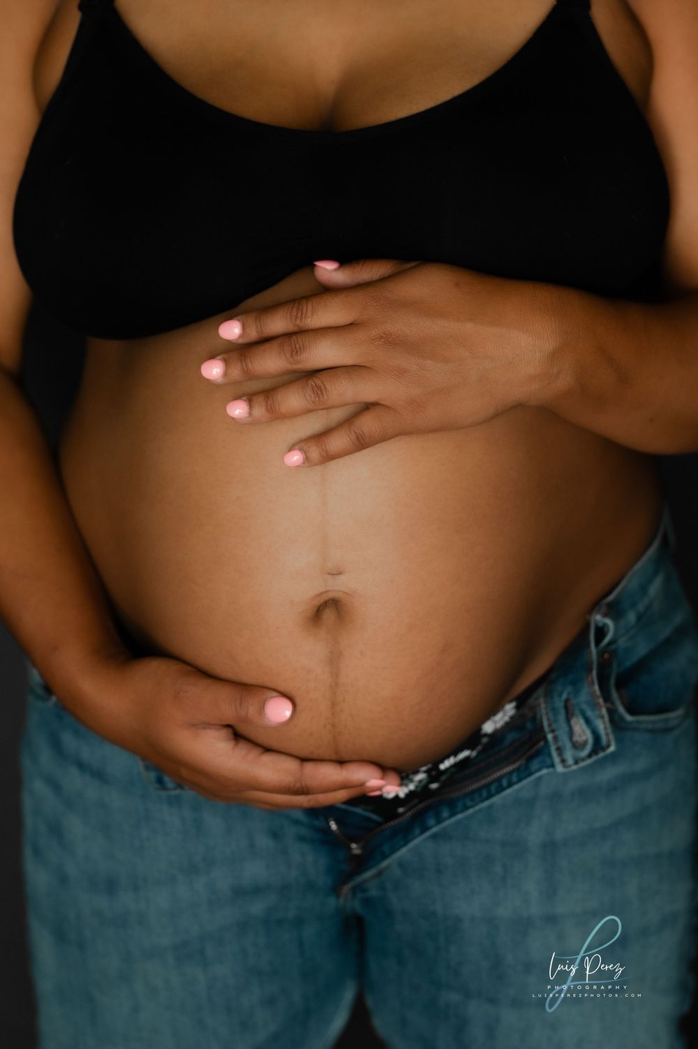 A pregnant person wearing a black top and jeans cradles their belly with manicured hands in an intimate maternity photo.